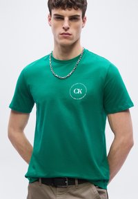 Νέος άνδρας που φοράει πράσινο t-shirt Calvin Klein με λευκό λογότυπο στο στήθος, αλυσίδα λαιμού από ασημί, και μπεζ παντελόνι σε απλό ανοιχτό φόντο.