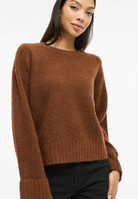 Pruun villane kampsun ümarate kaelusega, lahtise olekuga, soonikkoes varrukate ja äärega. Tekstureeritud kangas pehme välimusega, kombineeritud mustade pükstega.