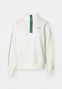 Krema krēpes sporta krekls ar augstu apkakli, zaļu rāvējslēdzēju un zaļām sānu svītrām. Iekļauta neliela rozā Nike logo uz krūšu daļas.