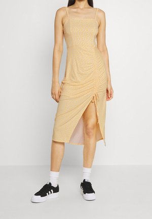 Mujer con un vestido midi amarillo con lunares blancos, abertura lateral fruncida, calcetas blancas y zapatillas negras, de pie frente a un fondo blanco.