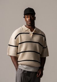 Polo din tricot bej cu mâneci scurte, model texturat, dungi orizontale negre și guler alb. Se poartă pantaloni gri și o șapcă neagră.