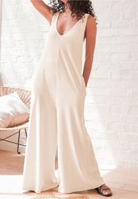 Beige mouwloze jumpsuit met een diepe V-hals, zijzakken en een brede broekspijp silhoët, gemaakt van zachte, vallende stof.