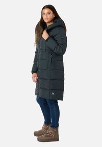 Lang, mørkegrøn pufferjakke med hætte, der har lynlås og store lommer samt vandret quiltet design. Bæres med blå jeans og brune støvler.
