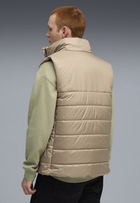 Beige puffervest magas gallérral, vízszintes steppelt panelekkel. Zöld textúrált hosszúujjú ing felett viselve, hátulról látható.