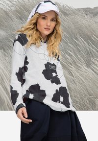 Witte mesh hoodie met zwart bloemenpatroon, met de tekst "COLD DREAMS." Gecombineerd met een marineblauwe rok, draagt het model een witte pet.
