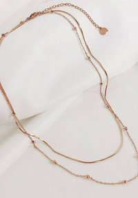 Collana a strati in oro rosa con catene delicate, accenti sferici e un piccolo ciondolo. Texture liscia con un design minimalista su sfondo chiaro.