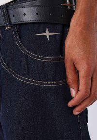 Pantalones vaqueros de mezclilla oscura con costuras doradas y un destacado adorno de estrella plateada. Incluyen un cinturón negro de cuero con una textura suave.