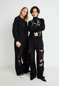 Sort ribbet zip-up sweater med sølv detaljer, parret med oversized sorte bukser med store slidte lapper. Sorte fodtøj.