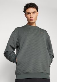 Sweatshirt en velours vert foncé avec un motif géométrique, des poignets et un ourlet côtelés. Présente un logo brodé subtil sur la poitrine.