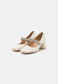 Scarpe bianche in pelle con tacco a blocco, punta quadrata e ampia cintura alla caviglia decorata con gemme trasparenti. Texture liscia e design minimal.