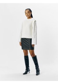 Maglione con corpo lavorato a maglia e maniche lunghe con pizzo; abbinato a una gonna di jeans nera e stivali neri con tacco al ginocchio.