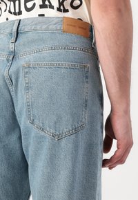 Blugi din denim albastru deschis, cu o croială lejeră. Prezintă un eticheta cusută pe talie și un singur buzunar pe spate cu o textură subtilă.