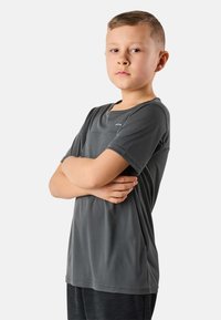Jongen met kort blond haar staat met gekruiste armen, draagt een donkergrijs T-shirt met korte mouwen en een zwarte broek, tegen een effen lichte achtergrond.