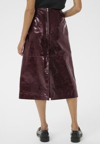 Le modèle se tient de dos par rapport à la caméra, portant une jupe midi bordeaux brillante et des chaussures noires. La jupe possède une fermeture éclair au dos.