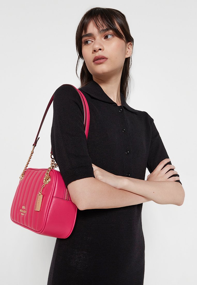 Rosa quiltad handväska med en guldkedjesladd och en tag. Texturerad yta, kompakt design. Bärs över axeln, kombinerad med en svart outfit.