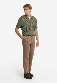 Ljus olivgrön polo skjorta med korta ärmar, ihop med beiga byxor. En stickad tröja är hängd över axlarna. Svarta skor.