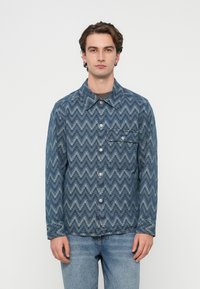 Jachetă din denim cu un model zigzag albastru, având guler, nasturi în față și un singur buzunar la piept cu închidere cu nasture.