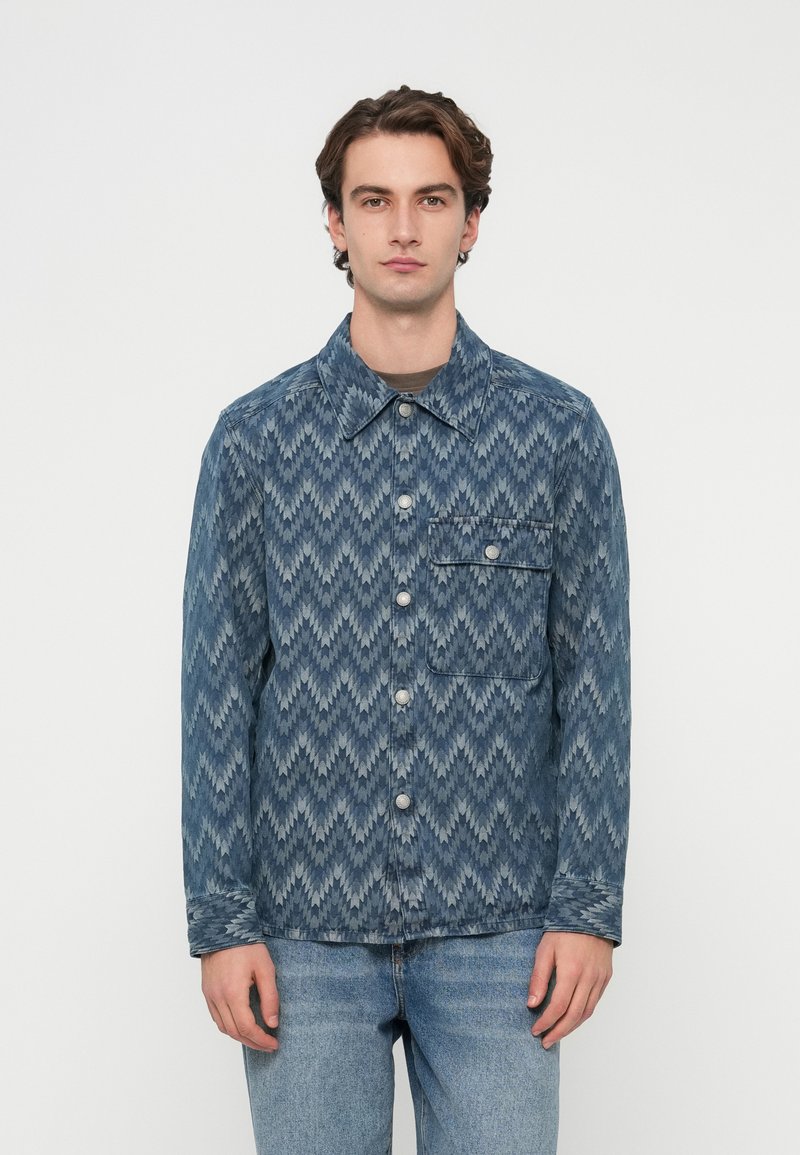 Jachetă din denim cu un model zigzag albastru, având guler, nasturi în față și un singur buzunar la piept cu închidere cu nasture.