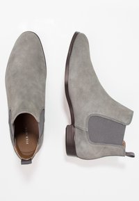 Bottines en daim gris avec panneaux latéraux élastiques et talon en bois sombre. Elles présentent un bout arrondi et une languette de traction à l'arrière.