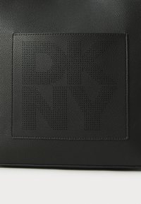 Black textured leather bag featuring a perforated rectangular logo patch with stylized letters "RKNY" in a bold, modern font.