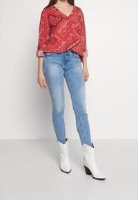 Blouse rouge à motifs avec un col en V, jean skinny bleu, et bottes de cow-boy blanches à bout pointu et talon bloc.