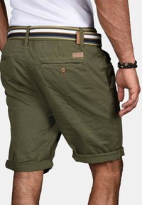 Mann trägt olivgrüne hochgekrempelte Shorts mit gestreiftem Gürtel und Perlenarmband am rechten Handgelenk, steht mit locker hängender rechter Hand.