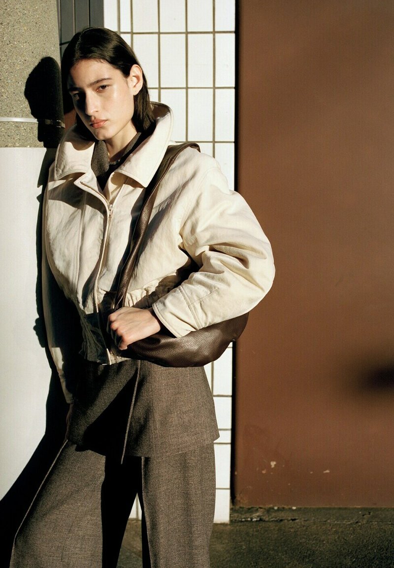 Jeune personne appuyée contre un mur carrelé, vêtue d'une veste ivoire zippée, d'un pantalon taille haute marron, et portant un sac à bandoulière marron foncé.