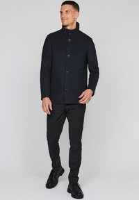Abrigo de mezcla de lana en azul marino con un cuello alto, que presenta seis botones grandes y un corte entallado. Combinado con pantalones negros ajustados y zapatos negros.