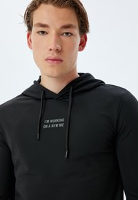 Sudadera con capucha negra hecha de tela suave. Cuenta con cordones y un texto impreso en el pecho que dice: "ESTOY TRABAJANDO EN UN NUEVO YO."
