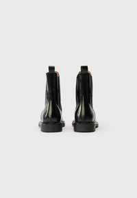 Kaksi kiiltäviä mustia nahkasaappaita nähtynä takaa, joissa on vetoketjut ja matala lohko- korko, yksinkertaisella valkoisella taustalla.