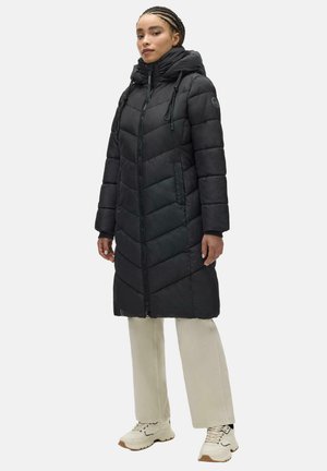 Kvinde iført en lang sort polstret hættejakke, beige bukser og hvide sneakers, stående foran en ensfarvet lys baggrund.