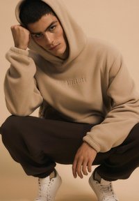 Fleece'owa bluza w kolorze taupe z haftowanym logo, luźny krój i regulowany kaptur, w połączeniu z ciemnymi dresami i białymi sneakersami.