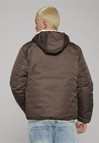 Brązowa kurtka puchowa z teksturowaną powierzchnią, pikowanym wzorem i kapturem. Kurtka ma elastyczne mankiety oraz kremową podszewkę kontrastującą z zewnętrzną częścią.