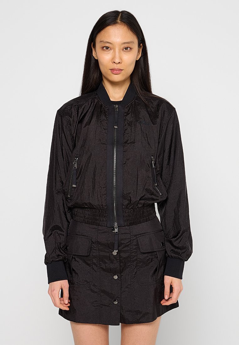 Schwarze Bomberjacke aus krinkligem Material, mit einem Reißverschluss vorne, zwei Reißverschlusstaschen und gerippten Bündchen. Kombiniert mit einem passenden Rock.