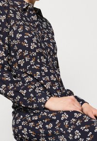 Blusa navy con motivo floreale in bianco e ruggine, dotata di colletto, chiusura con bottoni sul fronte e maniche lunghe. Tessuto morbido con una consistenza liscia.