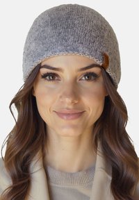 Kvinde med langt brunt hår iført en grå strikket hue og beige tøj, der smiler blidt med en neutral baggrund.