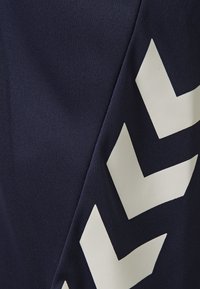 Marinblått tyg med en texturerad yta, prytt med stora vita chevron-mönster som är diagonalt placerade. Designen betonar moderna grafiska element.