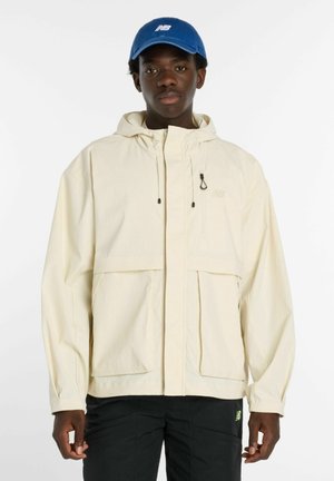 Jeune homme portant une veste à capuche crème, une casquette bleue avec un logo et un pantalon noir, debout devant un fond blanc uni.