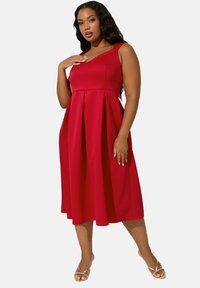 Punainen hihaton mekko, jossa on A-linjainen siluetti, istuva päälliosa ja laskostettu hame. Tasainen kangas, jota korostavat kultaiset sandaalit.