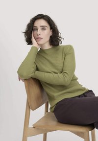 Rippstrukturierter, langärmliger Pullover in Olivgrün mit Rundhalsausschnitt. Das Model sitzt auf einem Holzstuhl und trägt braune Hosen, der Hintergrund ist in sanften Neutralton gehalten.
