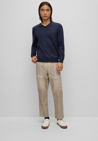 Ναυτικό πλεκτό πουλόβερ με V, φτιαγμένο από μαλακό υλικό, συνδυασμένο με μπεζ cargo παντελόνι με πλευρικές τσέπες. Λευκά sneakers με καφέ λεπτομέρειες στη σόλα.