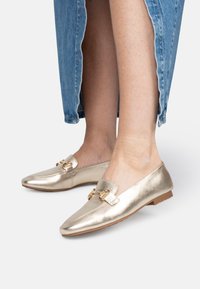 Metallische gouden loafers met een gladde textuur, voorzien van een decoratieve dubbele gesp op de bovenkant en een lage platte zool.