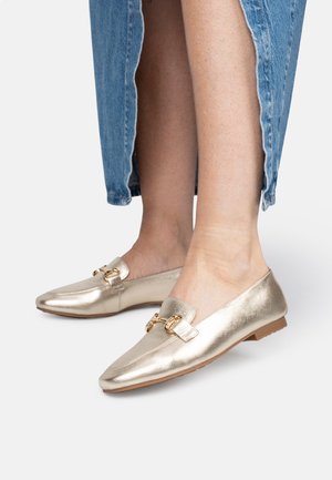 Des mocassins dorés métalliques avec une texture lisse, dotés d'une double boucle décorative sur le dessus et d'une semelle plate et basse.