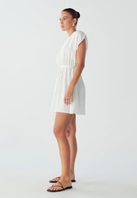 Weißes kurzes Kleid mit kurzen Ärmeln, Tunnelzug in der Taille und ausgestelltem Saum. Modell trägt schwarze Riemchensandalen und steht vor einem hellen Hintergrund.