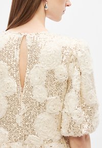Μπεζ φόρεμα με υφή υφ�άσματος, διακοσμημένο με παγιέτες και ανθικές εφαρμογές. Λεπτομέρεια με άνοιγμα στο πίσω μέρος και φουσκωτά μανίκια. Κομψός σχεδιασμός.