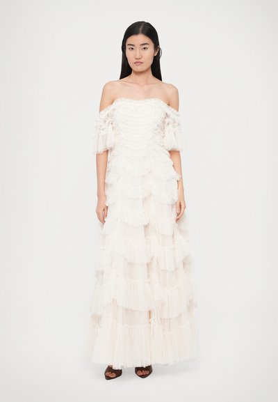 Off-the-shoulder, tiered gown in light cream with ruffled fabric and polka dots. Long sleeves and a flowing silhouette. Brown shoes below.