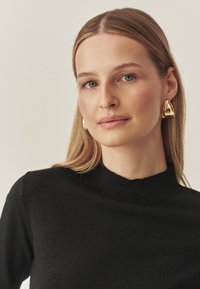 Goldene, dreieckige Ohrringe mit polierter Oberfläche, kombiniert mit einem schwarzen Rollkragenpullover aus strukturiertem Strickstoff. Neutraler Hintergrund.