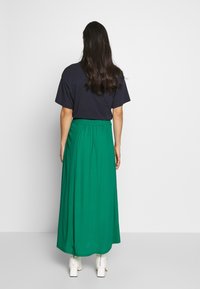 Groene maxi rok met een elastische tailleband, gecombineerd met een marineblauw T-shirt. De stof lijkt glad, met enkelhoge witte laarzen zichtbaar.