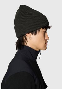 Dzianinowa ciemnozielona czapka z podwiniętym mankietem i fakturowanym wzorem, noszona przez osobę w czarnej kurtce z wysokim kołnierzem.