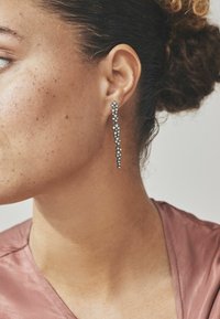 Orecchini a goccia in argento presentano un design a cascata con più pietre rotonde, che riflettono la luce, indossati all'orecchio sinistro accanto a una scollatura liscia.